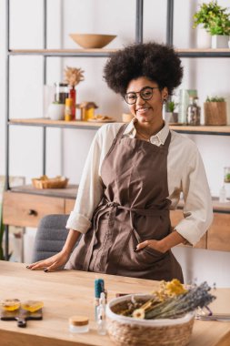 happy african american woman with hand in pocket of apron standing near table with blurred soap bars and dried herbs  clipart