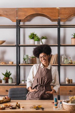 smiling african american woman standing with hands in pockets of apron near natural ingredients for soap and candle making