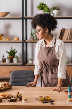 african american craftswoman in eyeglasses and apron looking at soap bars near natural ingredients on table