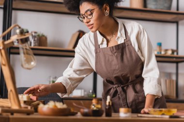 african american woman in apron and eyeglasses taking soap bars near natural ingredients on blurred foreground clipart