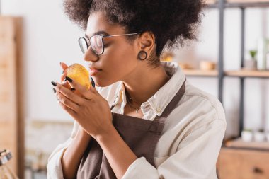 african american craftswoman in apron and eyeglasses smelling aromatic soap in blurred workshop
