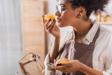 african american craftswoman in eyeglasses and apron smelling handmade soap in workshop