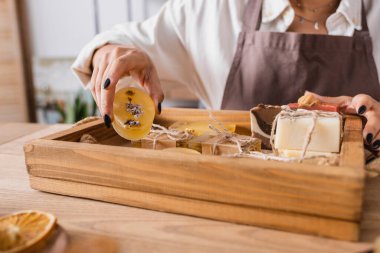 partial view of african american craftswoman showing bar of herbal soap near wooden tray in workshop