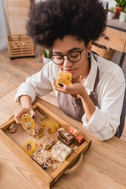 high angle view of african american craftswoman in eyeglasses enjoying scent of herbal soap in workshop