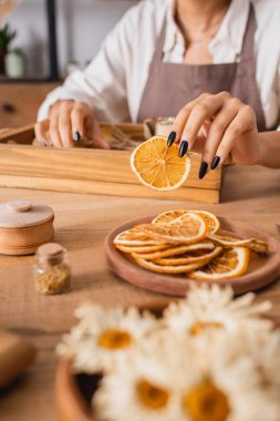 partial view of african american woman holding dried orange slice near camomiles on blurred foreground
