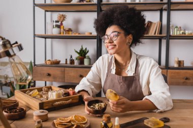 pleased african american craftswoman in eyeglasses holding bar of handmade soap and wooden bowl with dried herbs in workshop