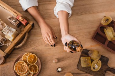 top view of cropped african american woman holding essential oil near homemade soap and dried orange slices in craft workshop clipart