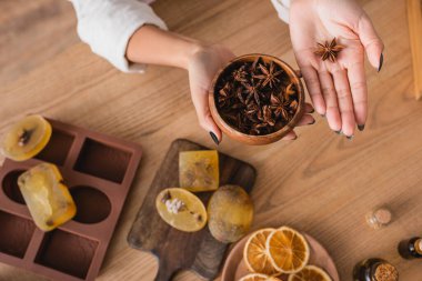 top view of cropped african american woman holding wooden bowl with aniseeds above blurred homemade soap and dried orange slices