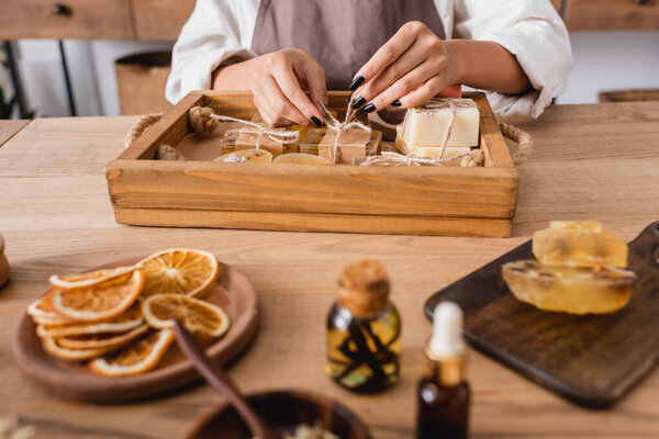 cropped view of african american craftswoman tying twine on handmade soap near dried orange slices and essential oils on blurred foreground