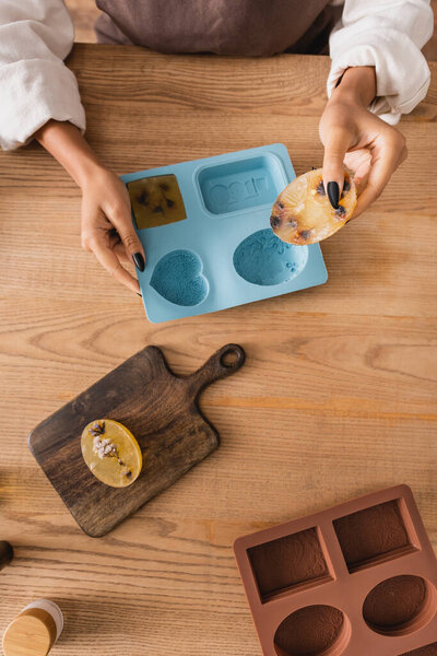 top view of cropped african american woman holding handmade soap near silicon molds and cutting board on wooden table
