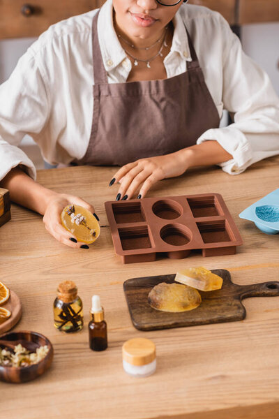 cropped view of african american craftswoman holding herbal soap near silicone molds and natural ingredients