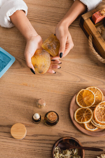 top view of cropped african american craftswoman holding homemade soap near dried orange slices and essential oils