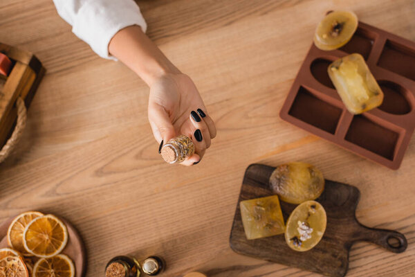 top view of cropped african american woman holding jar with dried herbs near soap bars on chopping board and silicone mold