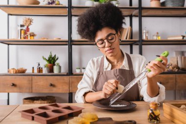 african american craftswoman in eyeglasses grating soap bar near silicone mold and essential oil