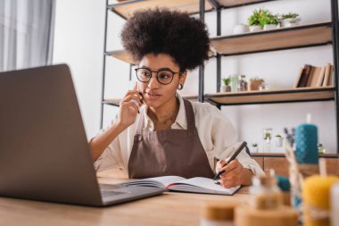 Not defteri ve kalem ile bilgisayarın yanındaki akıllı telefondan sipariş alan Afro-Amerikan bir sanatçı. 