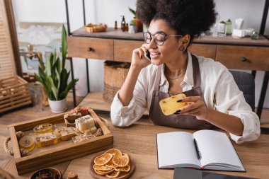 smiling african american craftswoman looking at homemade soap while talking on smartphone near natural ingredients  clipart