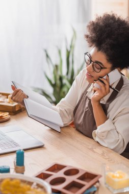 african american woman in apron and eyeglasses holding notebook and talking on cellphone near laptop in craft workshop clipart