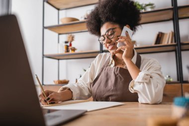 smiling african american craftswoman talking on smartphone and writing order in notebook near blurred laptop in workshop