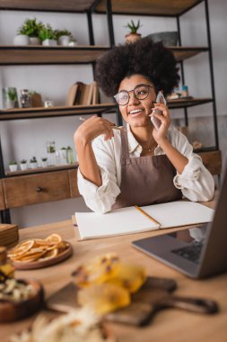 cheerful african american woman in eyeglasses and apron talking on cellphone near empty notebook and laptop in craft workshop