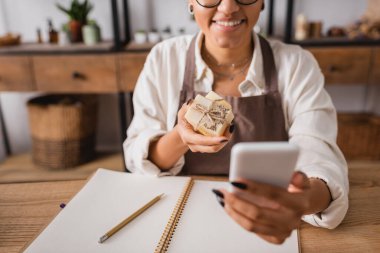 cropped view of positive african american craftswoman holding herbal soap during video call on smartphone near notebook