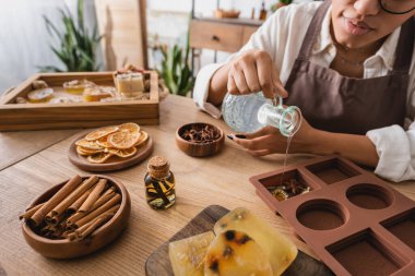 partial view of african american craftswoman pouring liquid soap in silicone mold near spices and cinnamon sticks with dried orange slices