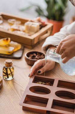 cropped view of african american woman pouring liquid soap in silicone mold with dried herbs near essential oil on wooden table