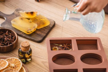 partial view of african american craftswoman pouring liquid soap in silicone mold with dried herbs near essential oil and orange slices