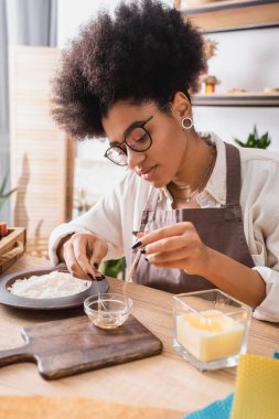 african american woman in eyeglasses dipping twine in melted wax near chopping board and burning candle clipart