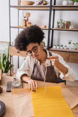 african american craftswoman in apron and eyeglasses holding candle wick near natural wax sheet and parchment on wooden table