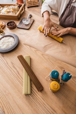 partial view of african american woman rolling wax sheet on parchment near homemade candles on wooden table