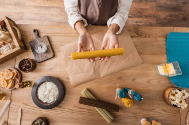 top view of cropped african american woman holding handmade candle near parchment and natural ingredients in craft workshop