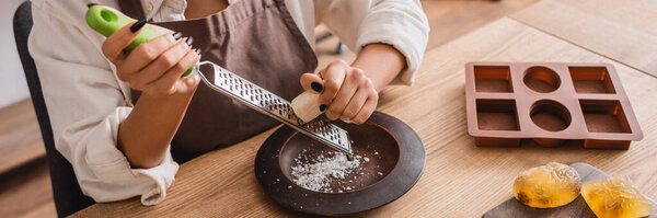 cropped view of african american woman grating soap near silicone mold on wooden table in workshop, banner