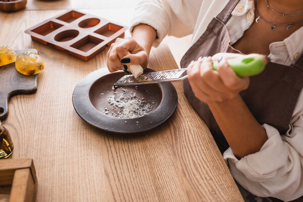 partial view of african american woman grating soap bar near wooden plate and silicone mold in craft workshop