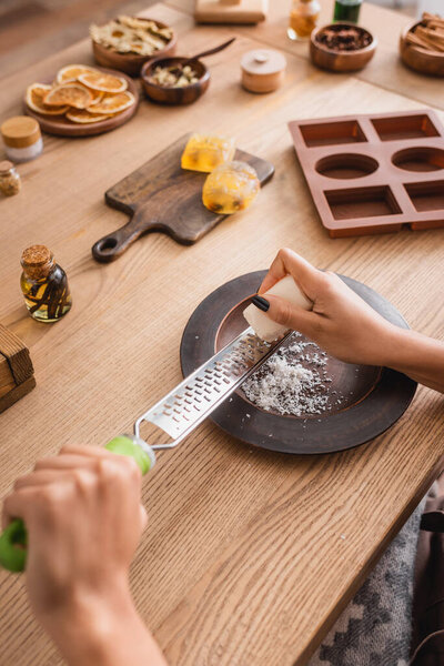 partial view of african american woman grating soap bar near natural ingredients and silicone mold on wooden table