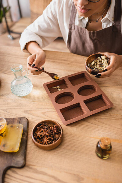 cropped view of african american woman holding wooden bowl with dried herbs near silicone mold and pitcher with liquid soap