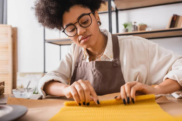 Stock image african american craftswoman in apron and eyeglasses rolling wax sheet on parchment while making candle in workshop