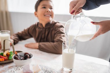 Muslim mother pouring milk during suhur breakfast near blurred son at home 