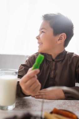 Side view of cheerful muslim boy holding blurred cevizli sucuk during suhur at home 