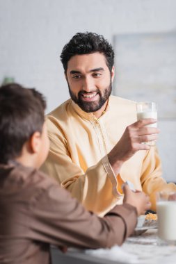 Gülümseyen Müslüman baba elinde bir bardak süt tutuyor ve sabah bulanık oğluyla konuşuyor. 