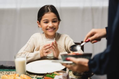 Smiling muslim girl holding glass of milk near blurred mom and ramadan dinner  clipart