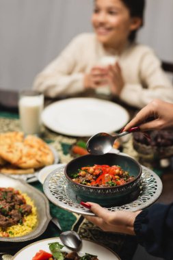 Muslim woman holding tasty dish near blurred daughter and ramadan dinner  clipart