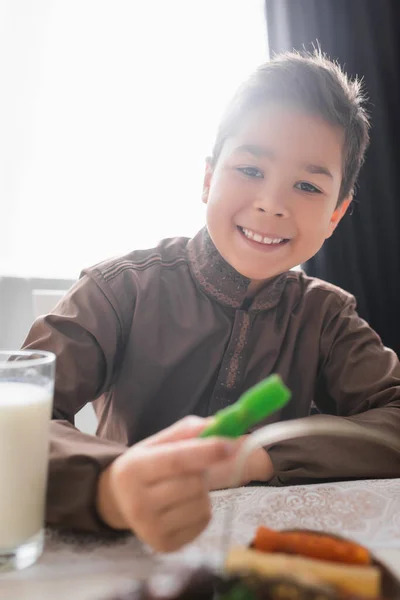 Smiling muslim boy holding blurred cevizli sucuk near glass of milk during ramadan breakfast