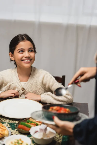 Smiling muslim girl looking at blurred mom with food during ramadan dinner at home