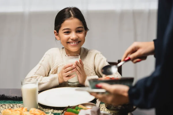 Smiling muslim girl holding glass of milk near blurred mom and ramadan dinner