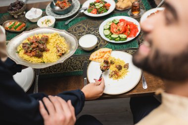 Muslim woman putting pilaf on plate near blurred husband at home  clipart