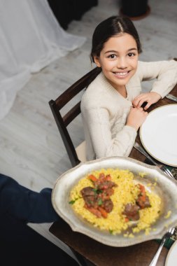 High angle view of smiling muslim girl looking at camera near blurred mom with iftar pilaf at home  clipart