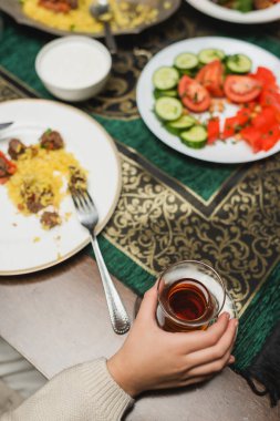 Top view of muslim girl holding turkish tea glass near blurred ramadan dinner at home  clipart