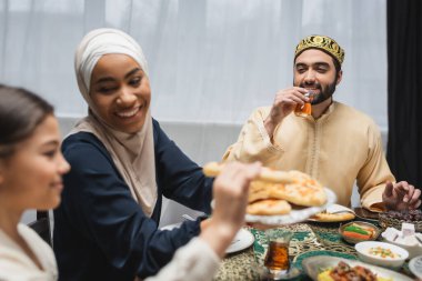 Smiling muslim man holding turkish tea glass near family and ramadan dinner  clipart