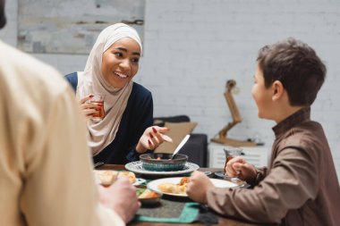 African american woman in hijab holding tea and talking to son near husband and food at home  clipart