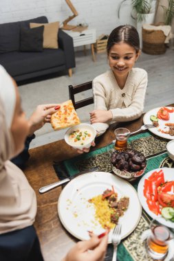 High angle view of smiling muslim girl holding dish near blurred mother during ramadan dinner at home 